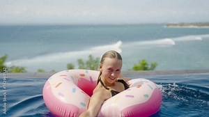 Young woman emerging from inflatable rubber ring in water with arms in infinity ocean view pool on luxury private villa. Happy memories of summer vacation, smile swim and roll, have fun on pool party