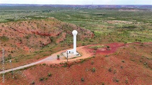 The white weather radar Tower sits beside a small utility complex in arid red-soil landscape. Distant Mount Isa City and sparse vegetation create a contrast between man-made infrastructure and nature