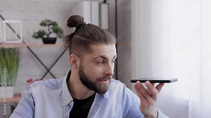Smiling young man recording voice message on smartphone. Handsome guy using virtual assistant app sitting at home. Voice recognition, audio messages, using translator on mobile phone.