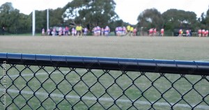 A mesh metal barrier at a sports ground, with a blurry view of two groups of Australian football players finishing games and dispersing. concept of school activity, with high school students competing