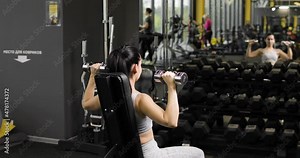 Young brunette woman doing overhead presses with dumbbells. She trains her shoulders, doing an overhead press sitting on a bench in the gym in front of mirror. Concept of sport and healthy lifestyle.