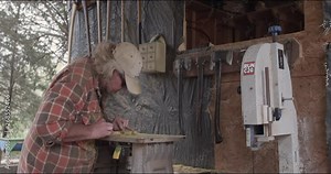 Carpenter runs hedge wood through a routering table as camera tilts up in 4K. Medium shot.