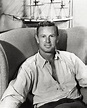 a man sitting on a couch with a model ship in the back ground behind him