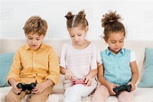 Adorable multiethnic kids sitting on couch and playing with joysticks ...