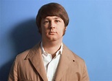 a man in a tan jacket and white shirt is standing against a blue wall ...