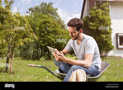 Man Sitting In Wheelbarrow In Garden Using Digital Tablet Stock Photo