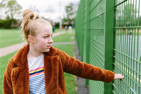 Preteen Girl Looking Through The Railings In A Park Enclosing Sports Pitches By Stocksy