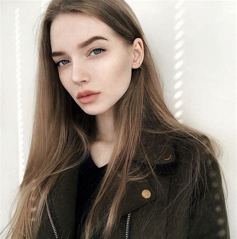 a woman with long brown hair wearing a black jacket and standing in front of a white wall