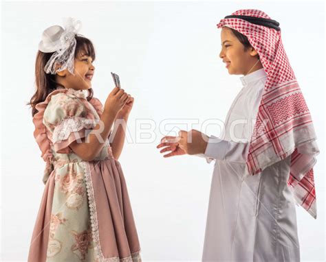 خلفية بيضاء لصبي وفتاه صغيران سعوديان مبتسمان ، تمسك الفتاه الصغيرة العيدية بيدها ينظر الطفل