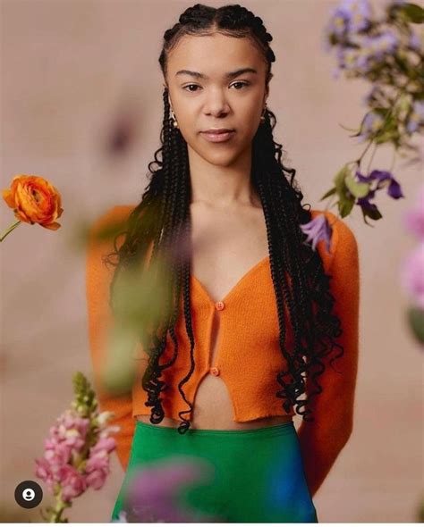 a woman with long braids standing in front of purple and orange flowers wearing an orange top
