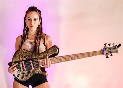 A Woman With Dreadlocks Is Holding A Guitar And Posing For A Photo In Front Of A Pink Background