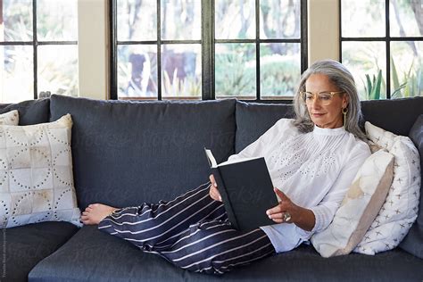 Mature Woman With Grey Hair Reading A Book On Sofa In Living Room By Stocksy Contributor