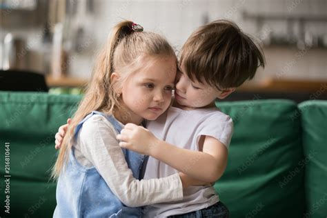Little Boy Hugging Consoling Upset Girl Sitting On Sofa Kid Brother