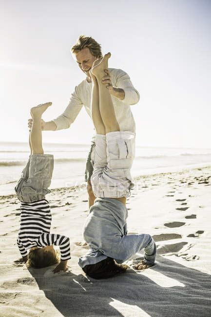 Father Helping Sons With Handstand On Beach — Rear View Love Stock