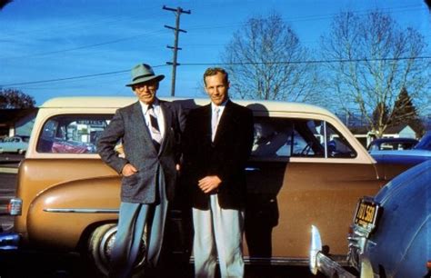 Two Men Standing Next To Each Other In Front Of An Old Car With Another Man Wearing A Suit And Tie