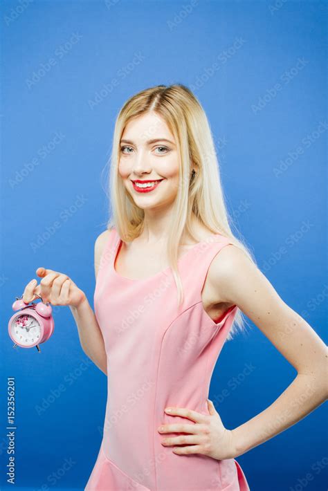 beautiful smiling blonde haired girl wearing dress with pink alarm clock in hand is posing on