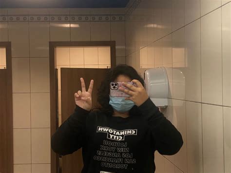 A Woman Wearing A Face Mask Taking A Selfie In A Public Restroom With