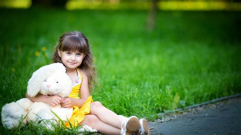 Cute Little Girl Is Wearing Yellow Dress Sitting On Green Grass In Blur