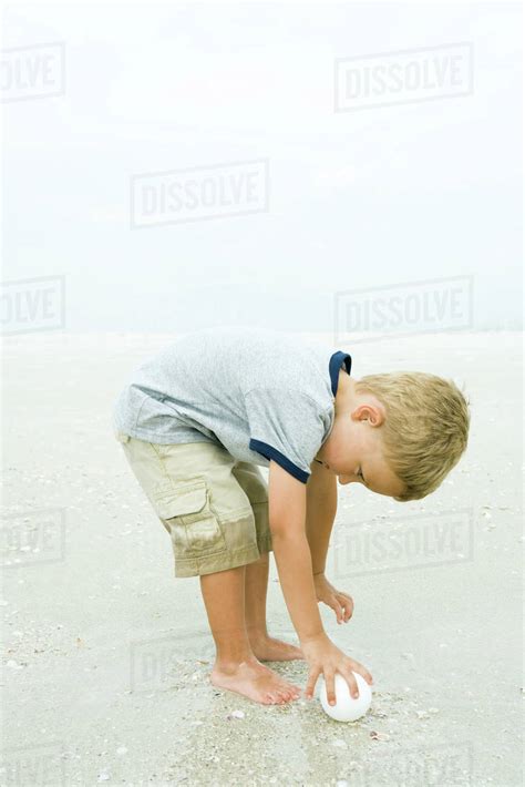 Little Boy Bending Over To Pick Up Ball On Beach Full Length Stock