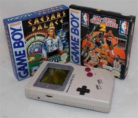 An Old Nintendo Gameboy Next To Two Other Games In Boxes On A White Surface