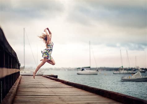 Young Girl Leaping On A Dock Above A River On A Cloudy Day By Angela