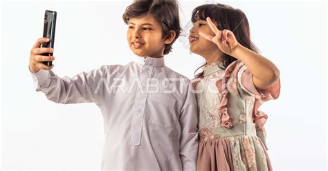 خلفية بيضاء لصبي وفتاه صغيران سعوديان مبتسمان ، يمسك الصبي الجوال بيده يقومان بإلتقاط صور