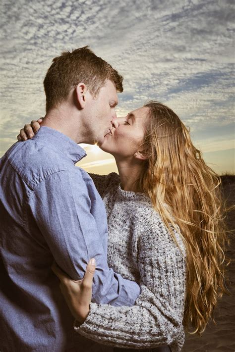 A Man And Woman Kissing Each Other In Front Of A Cloudy Sky With The Sun Setting Behind Them