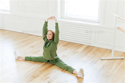 Strong Flexible Curly Red Haired Woman Does Side Leg Split Raises Hands Wants To Have Perfect
