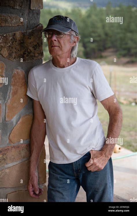 a youthful handsome 79 year old man in a white t shirt and jeans leans against a stone wall