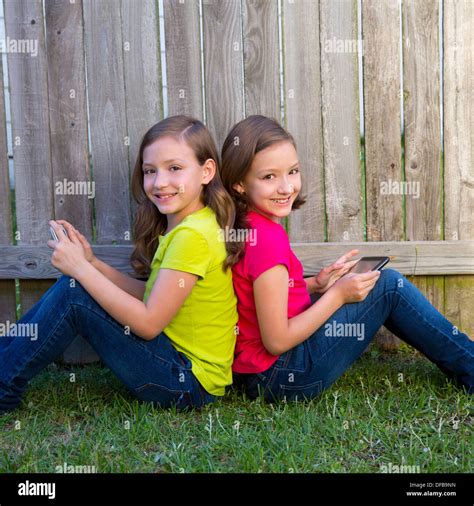 Twin Sister Girls Playing With Tablet Pc Sitting On Backyard Lawn Fence