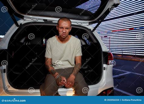A Dark Skinned Tattooed Guy In A White Shirt Sitting In An Opened Car