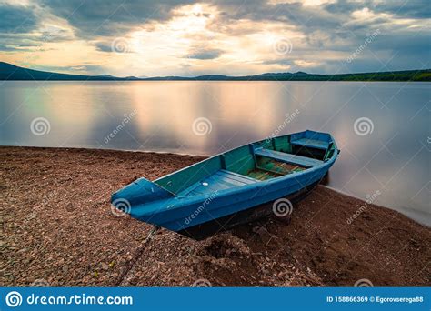 Blue Wooden Boat Moored On The Shore Of Lake Under Cloudy Sky At Sunset