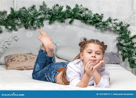 Little Cute Beautiful Girl Lies On Her Stomach On The Bed In Blue Jeans