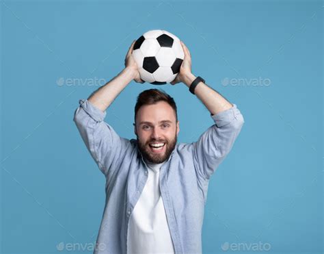 Smiling Young Man Holding Soccer Ball Above His Head On Blue Studio