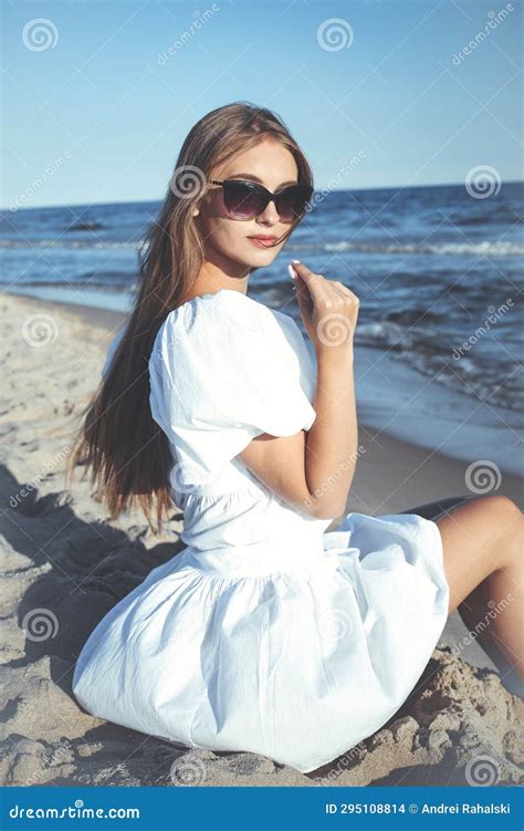 Happy Beautiful Blonde Woman Is Sitting On The Ocean Beach In A White Summer Dress And