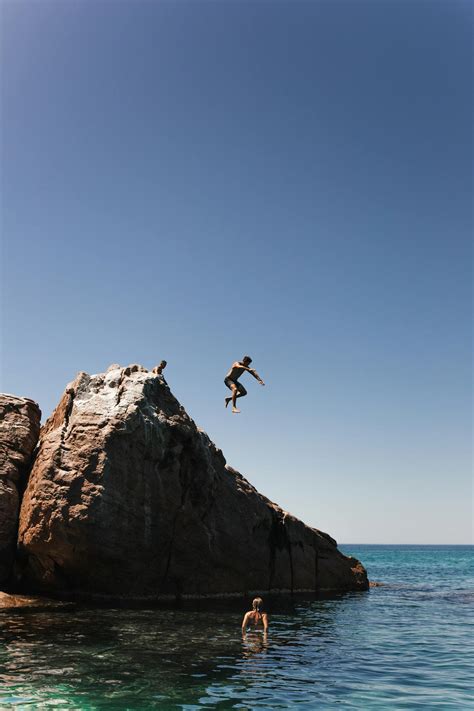 Unrecognizable Man Jumping Off Cliff Into Clear Blue Water On Summer