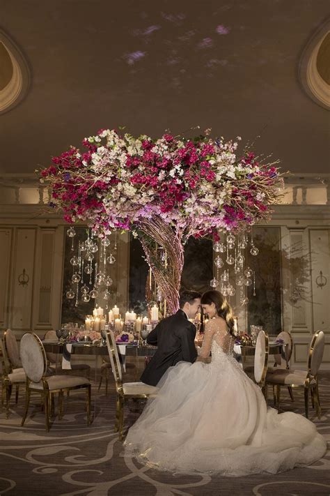 A Bride And Groom Sitting At A Table With Flowers In The Center
