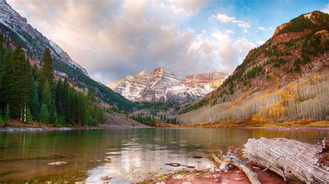 A Lake Surrounded By Mountains And Trees With Rocks In The Foreground