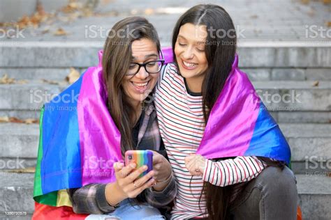 beautiful lesbian couple with rainbow flag using a mobile phone in the street high quality photo