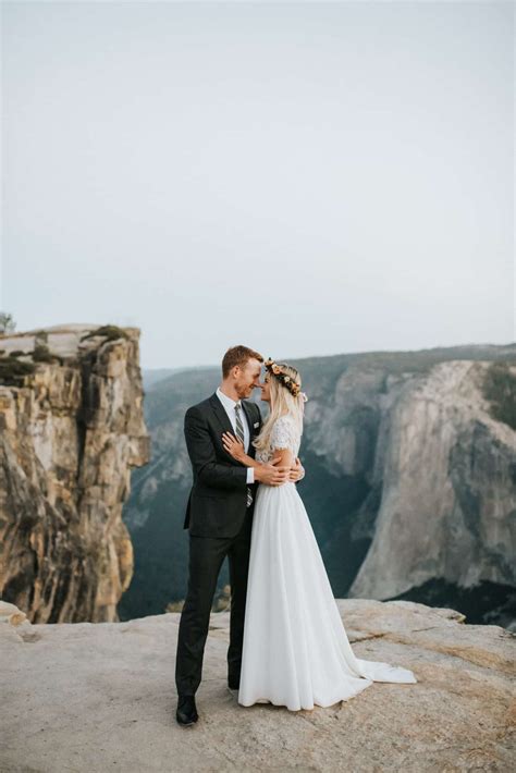 A Bride And Groom Standing On Top Of A Cliff In Front Of The Mountains
