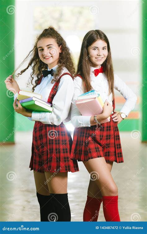 Two Schoolgirls In Red School Checkered Uniforms Stand In A Park Near A Aaa
