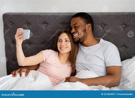 Millennial Multiracial Couple With Smartphone Taking Selfie In Bed Before Going To Sleep