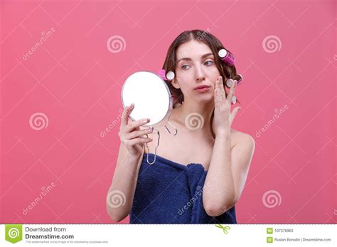 A Girl Holds A Mirror In Hands And Looks Closely At It Touching Her Face On A Pink Background