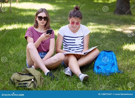 De Foto Van Twee Vrouwelijke Tieners Zit Op Groen Gras Openlucht Las Boek En Gebruik Mobiele