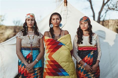 Three Women Standing Next To Each Other In Front Of A Teepee With A Colorful Quilt On It