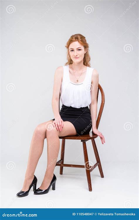 Young Beautiful Woman With Long Red Curly Hair Sitting On A Wooden Chair On A White Background