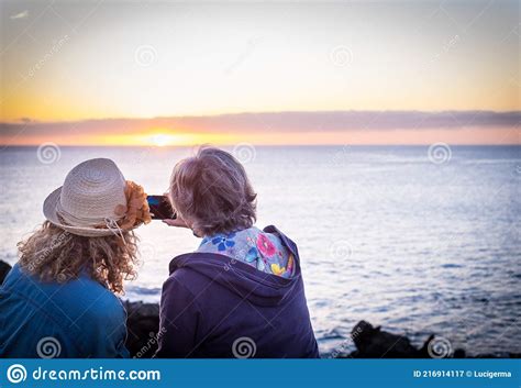 Two Women Sitting Admiring The Sunset Over The Sea While Taking A Photograph With Their Mobile