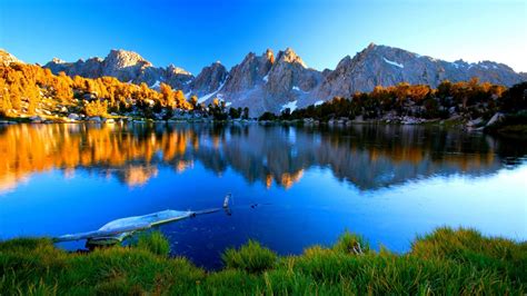 Summer Mountain Lake Shore With Green Grass Pine Trees Mountains Rocky Peaks With Snow