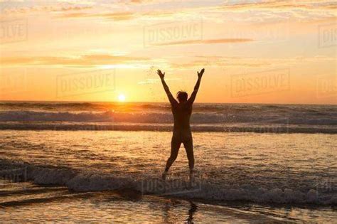 Rear View Of Mid Adult Nude Womans Silhouette Standing In Ocean At Sunset Arms Raised Stock