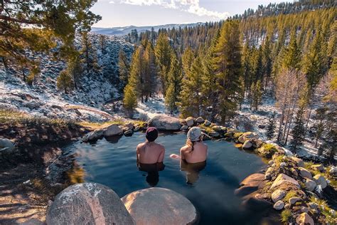 two people sitting in the middle of a pool surrounded by rocks and pine trees with snow on the